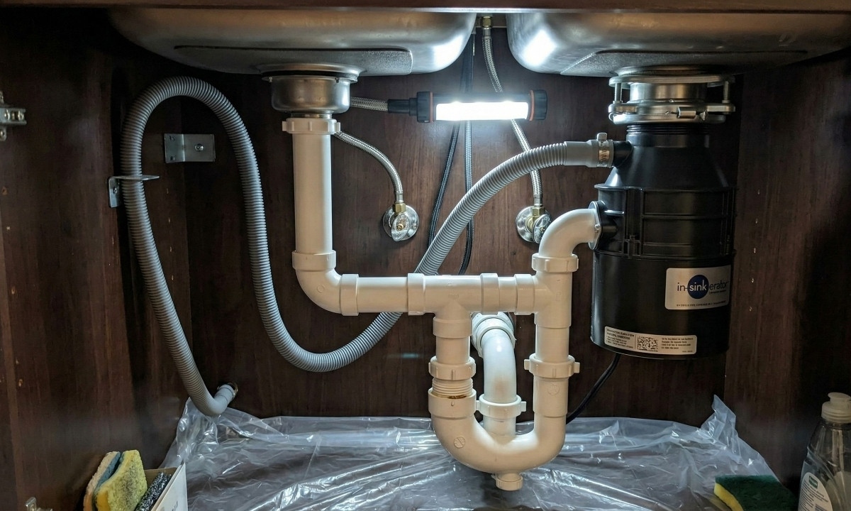 Under-sink view showing the complex drain configuration of a double-bowl sink with an attached garbage disposal and shared P-trap.